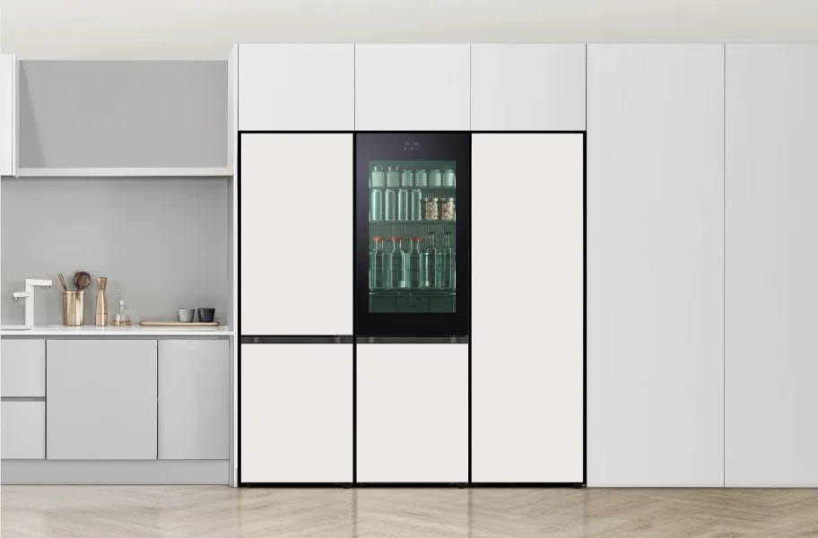 주방 공간에 설치된 대형 냉장고가 보입니다. 냉장고는 깔끔한 디자인으로, 중앙에 투명한 문을 통해 내부가 보입니다. 내부에는 여러 개의 병과 용기가 정리되어 있습니다. 주방 조리대에는 주전자와 컵이 놓여 있으며, 벽면은 밝은 색상으로 마감되어 있습니다. 바닥은 나무 재질로 되어 있습니다.
