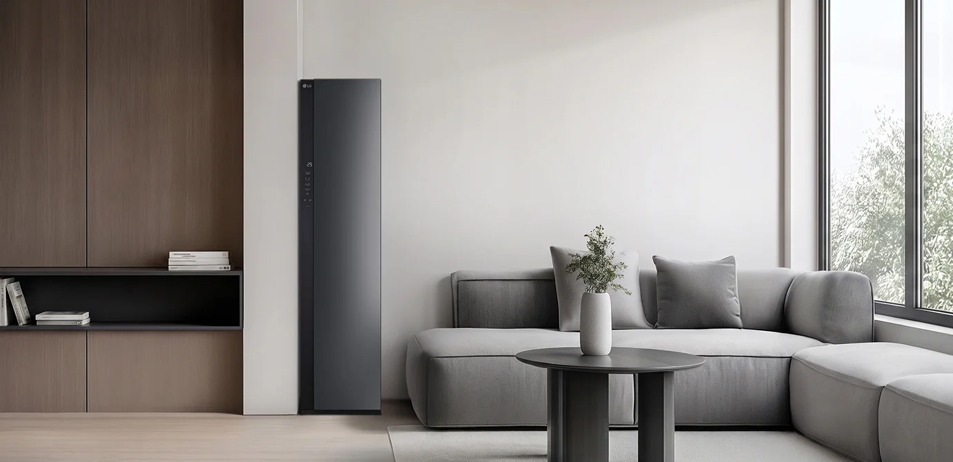 LG 스타일러 제품이 방의 왼쪽에 배치되어 있으며, 슬림한 디자인과 어두운 색상의 전면 패널이 특징입니다. 스타일러 옆에는 회색 소파가 놓여 있고, 소파 위에는 회색 쿠션이 있습니다. 소파 앞에는 작은 원형 테이블이 있으며, 테이블 위에는 흰색 꽃병과 꽃이 놓여 있습니다. 방의 오른쪽에는 큰 창문이 있어 자연광이 들어오고 있습니다.